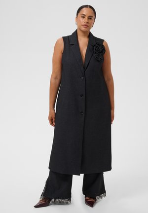Femme portant un long gilet noir sans manches avec un détail floral sur le revers, un pantalon noir à jambes larges avec un bord en dentelle, et des chaussures foncées pointues.