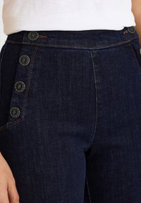 Pantalones de mezclilla oscura de talle alto con cierre lateral de botones, con detalles de botones en bronce y costuras contrastantes. Textura lisa y forma ajustada.