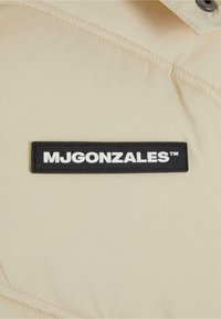 Beige jacket with a smooth texture. Features a black rectangular patch with the text "MJGONZALES™" in white. Minimalist design.