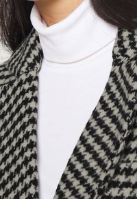 Abrigo de patrón de houndstooth en blanco y negro sobre un suéter de cuello alto blanco. El abrigo presenta una tela texturizada y un diseño de solapas.