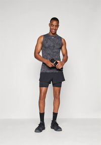 Rankovi tamsiai pilkos kamufliažo spalvos berankovis marškinėlis derinamas su juodais šortais. Modelis laiko vandens butelį, dėvi juodus sportinius batelius ir kojines.