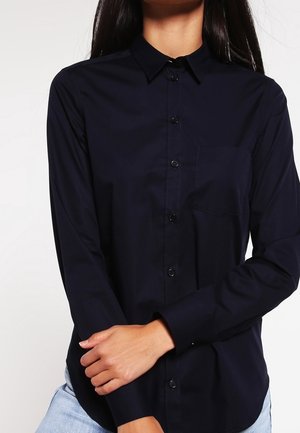 Person iført en langærmet sort skjorte med knapper, krave og brystlomme, kombineret med lyseblå jeans, armene krydset foran livet.