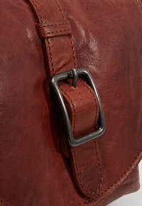 Detail van een bruine leren tas met een gestructureerde afwerking, een stevige metalen gesp en contrasterende stiksels langs de band en randen.