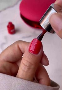Rode nagellak wordt aangebracht op een vingernagel, met een glanzende afwerking. Een klein rood flesje is zichtbaar op de achtergrond, en er ligt een ronde roze compact in de buurt.