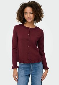Burgunderfärgad kofta i mjukt tyg med rund halsringning, knappstängning och volangärmar samt volangkant, tillsammans med blå jeans.