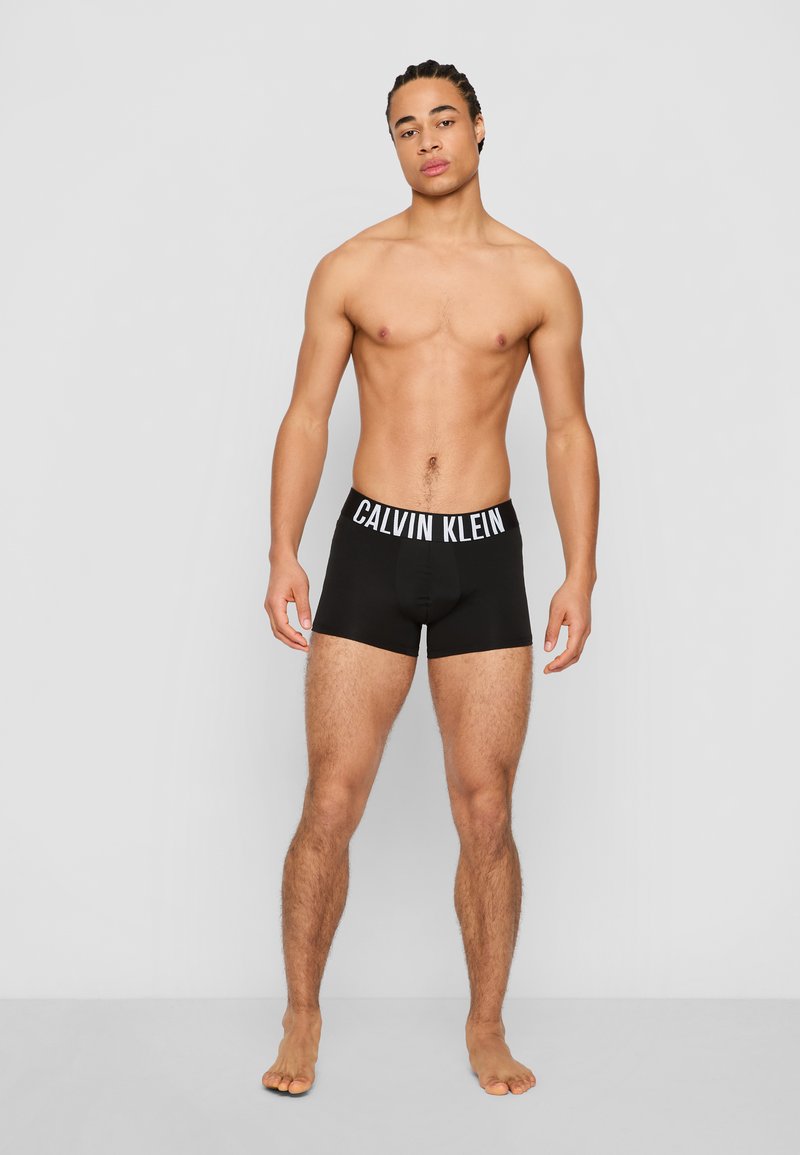 Mannelijk model met gevlochten haar staat blootsvoets tegen een effen achtergrond, gekleed in zwarte Calvin Klein boxershorts.