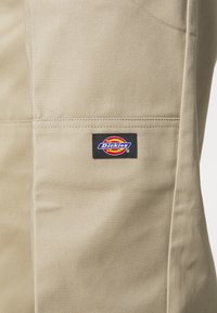 Beige Arbeitshosen mit glatter Textur, ausgestattet mit einem schwarzen und mehrfarbigen "Dickies"-Logo-Patch an der Seite und flachem Taschen-Design.