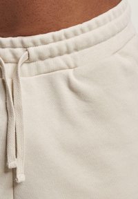 Beige puuvillaiset verkkarit, joissa on elastinen vyötärö, narut ja sivutaskut, esittelevät pehmeää rakennetta ja rentoa muotoilua.