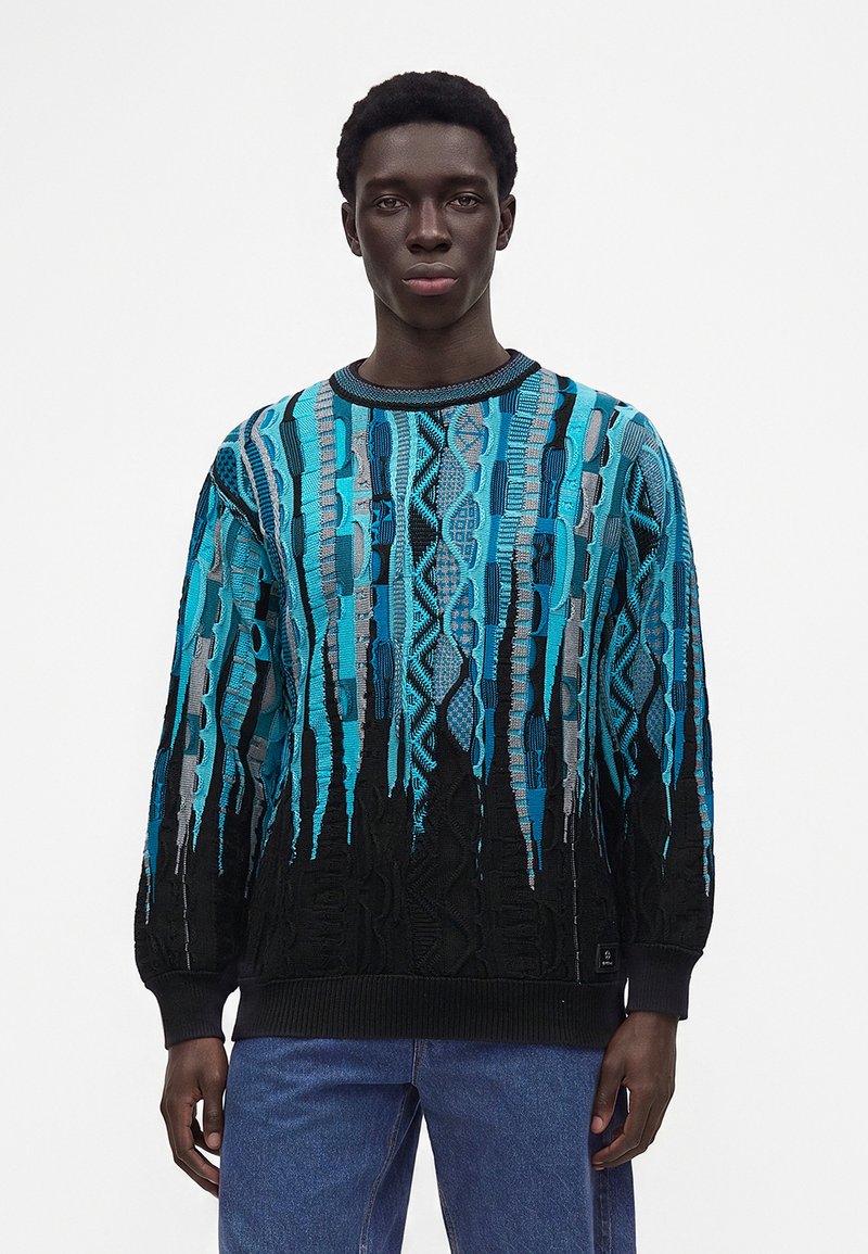 Homme portant un pull tricoté bleu et noir à motifs avec des rayures verticales texturées et un jean bleu, debout sur un fond blanc uni.