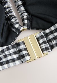 Sort stof med ternede hvide og sorte accenter, med en gylden metalspænde og rynket tekstur ved stropperne.