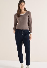 Pull marron à col en V associé à un pantalon de jogging bleu marine avec chevilles ajustées et deux poches latérales. Le mannequin porte des baskets beige.