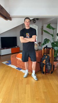Άνδρας με μαύρο μπλουζάκι και σορτς στέκεται με σταυρωμένα τα χέρια σε ένα σαλόνι κοντά σε καφέ καναπέ, φυτά εσωτερικού χώρου και ένα διπλωμένο μηχάνημα κωπηλασίας.