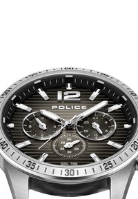Strieborné nerezové hodinky s pruhovaným čiernym ciferníkom. Obsahujú hodinové indexy, subciferníky pre dvojité časové pásma a výrazné "12" na vrchu.