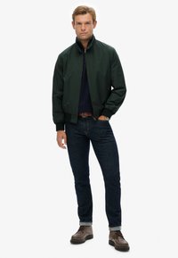 Donkergroene bomberjack met een hoge kraag, ritssluiting, ribgebreide manchetten, gedragen over een marineblauwe trui, in combinatie met donkere jeans en bruine laarzen.