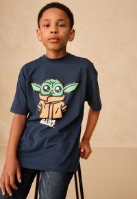 Navy t-shirt featuring a large green character graphic with round eyes and a brown coat, made of soft cotton with short sleeves.