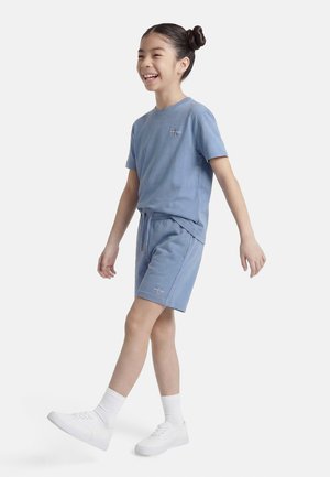 Niña joven con moños en el cabello, vestida con camiseta azul y pantalones cortos, calcetines blancos y zapatillas, caminando y sonriendo sobre un fondo blanco liso.