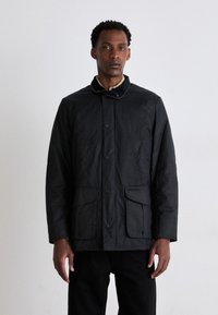 Veste noire, résistante à l'eau, avec un devant à boutons, de grandes poches à rabat et un col en velours côtelé, présentant une finition texturée.