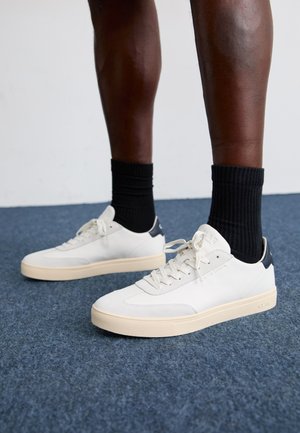 Valged nahast ja sametist spordijalatseid kreemika tallaga ning tumesinise kandiga, millel on lapikud nööbid ja tekstuuriga mustad sokid sinisel vaibal.