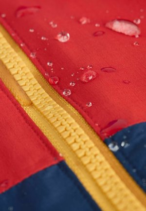 Vedenpitävä takki, jossa on punaisia, keltaisia ja sinisiä paneeleja. Tyylissä on näkyvä keltainen vetoketju ja selkeitä vesipisaroita kankaassa.