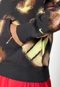 Nike hettegenser med et flerfarget abstrakt mønster; har en svart base, myk stoff, frontlomme, og ribbestrikkede ermer og nederkant.