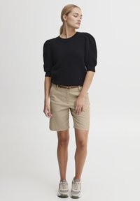 Svart teksturert genser med pufferm, kombinert med beige shorts og lyse joggesko. Brun belte fremhever antrekket.