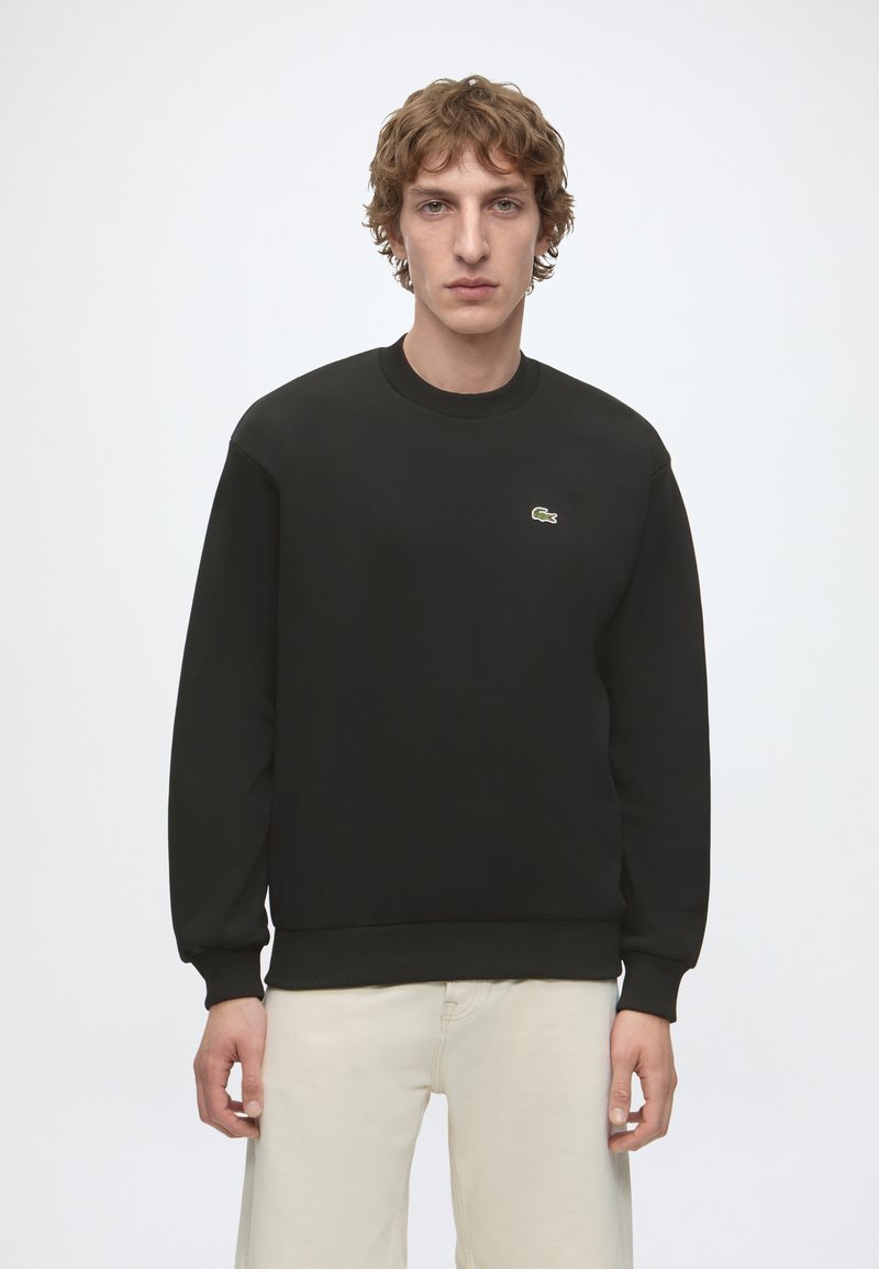 Jeune homme aux cheveux bouclés portant un sweat-shirt noir à manches longues avec un petit logo vert sur la poitrine et un pantalon clair.