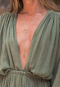 Femme portant une robe verte texturée à décolleté plongeant en V, taille froncée, et un collier lariat argenté avec une boucle circulaire et une barre pendante.