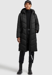 Abrigo de plumas negro con diseño largo, textura acolchada, capucha y bolsillos laterales. Combinado con un jersey negro ajustado, vaqueros oscuros y botas negras.