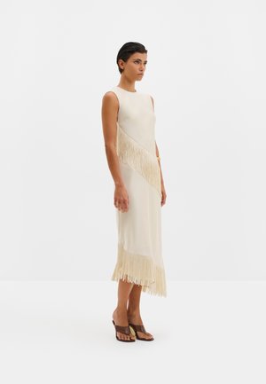 Femme portant une robe midi crème sans manches avec des détails en diagonale et des franges à l'ourlet, associée à des sandales à talons marron, se tenant de profil.