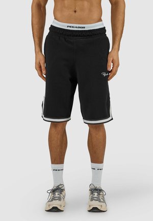 Mann trägt schwarze knielange Shorts mit weißer Zierlinie, weiße Socken, beige und graue Sneakers, steht vor einem schlichten Hintergrund.