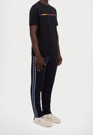 Man met een zwart T-shirt met een kleurrijke streep, zwarte broek met blauwe zijstrepen en witte sneakers, staand op een witte achtergrond.