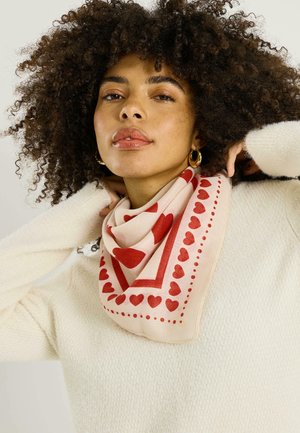 Mujer con cabello rizado que lleva un suéter blanco y una bufanda beige con estampado de corazones y puntos rojos, ajustándose la bufanda alrededor del cuello.