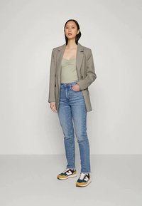 Mulher vestindo um blazer cinza, uma blusa verde-clara, jeans azuis e ténis multicoloridos, de pé com uma das mãos no bolso, contra um fundo liso.