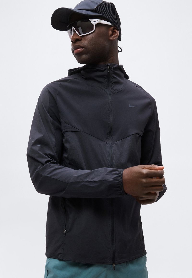 Hombre vestido con chaqueta cortavientos negra de Nike, gorra negra y gafas de sol deportivas blancas de Nike, ajustándose la manga con un fondo gris neutro.