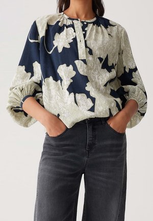 Vrouw draagt een losse bloes met grote witte bloemen op marineblauw, gecombineerd met hoog getailleerde donkergrijze jeans, handen in de zakken.