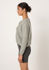 Moteris su garbanotais plaukais surištais kuodeliu, vilkinti šviesiai pilką pūstą megztinį ir tamsiai pilkus aptemptus šortus, stovinti šonu priešais vienspalvį foną.