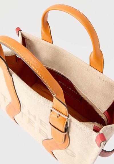 Μπεζ τσάντα χειρός από καμβά με δερμάτινες λαβές σε καφέ απόχρωση, με αγκράφες και εσωτερική τσέπη, με την ετικέτα "MARC JACOBS" στο εσωτερικό.