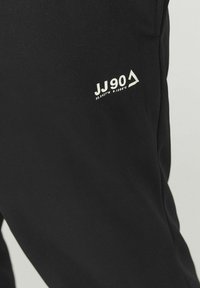 Pantalon noir en tissu lisse avec un logo blanc "JJ90" et des coordonnées. Design simple sans motifs ou accents supplémentaires.