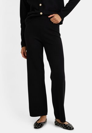 Pantalones de pierna ancha negros, hechos de una tela suave, con una cinturilla elástica, combinados con zapatos planos negros decorados con tachuelas plateadas.