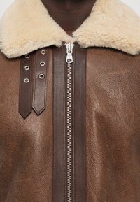 Veste en cuir brun avec un col en shearling beige, une fermeture éclair argentée et des sangles en cuir décoratives avec des œillets sur le côté gauche.