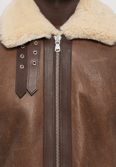 Braune Lederjacke mit einem beigen Schafslederkragen, einem silbernen Reißverschluss und dekorativen Lederbändern mit Ösen auf der linken Seite.