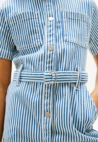 Camicetta di denim a righe con maniche corte, caratterizzata da righe verticali blu e bianche, due tasche sul petto, una chiusura con bottoni e una cintura in tessuto.