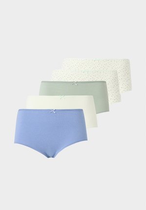 Femme aux longs cheveux foncés portant un soutien-gorge en dentelle blanche et un slip blanc taille haute assorti sur un fond clair uni.