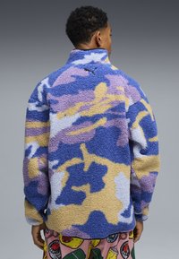 Giacca in pile con motivo mimetico blu, viola e beige, collo alto e logo nero sul retro. Superficie testurizzata e soffice.