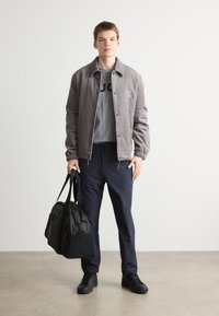 Grijs corduroy jack met knopen, grijze t-shirt met logo, marineblauwe broek, zwarte sneakers, een zwarte sporttas dragend, staand op een crème vloer.