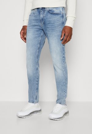 Jeans de denim azul claro con un diseño clásico de cinco bolsillos. Corte ajustado, textura ligeramente deslavada y cierre con cremallera. Llevados con zapatillas blancas.