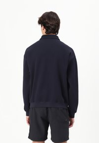 Sudadera azul marino con cuello tipo polo y puños acanalados, que presenta un corte relajado y una textura suave, combinada con pantalones cortos negros.