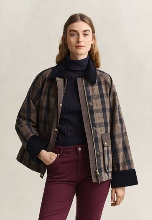 Donna con capelli mossi indossa una giacca a quadri marrone e nera sopra un maglione nero a collo alto e pantaloni bordeaux, in piedi con una mano in tasca.
