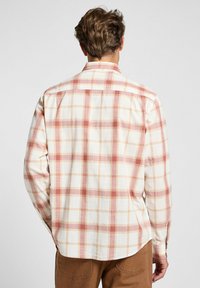Camisa de manga larga de cuadros en crema con cuadros rojos y beige. Presenta un collar con botones y un dobladillo curvado. Está hecha de un tejido ligero.