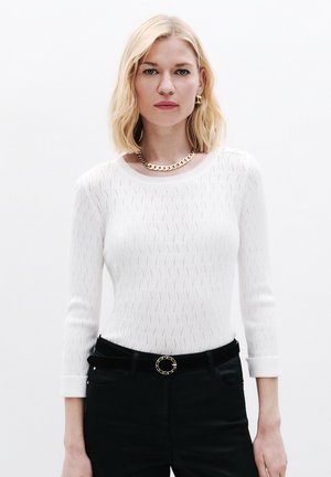 Weißer Strickpullover mit Wellenmuster, Rundhalsausschnitt und Dreiviertelärmeln, kombiniert mit schwarzen Hosen und einem goldenen Kettengürtel.